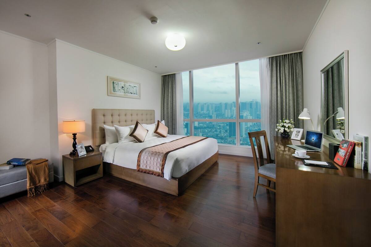 Een hotelkamer met een groot bed, een bureau met een laptop, een stoel en uitzicht op een stadsbeeld door grote ramen.