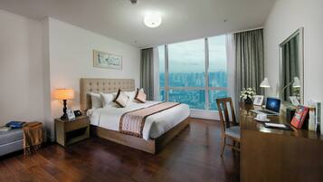 A hotel room with a large bed, a desk with a laptop, a chair, and a view of a cityscape through large windows.