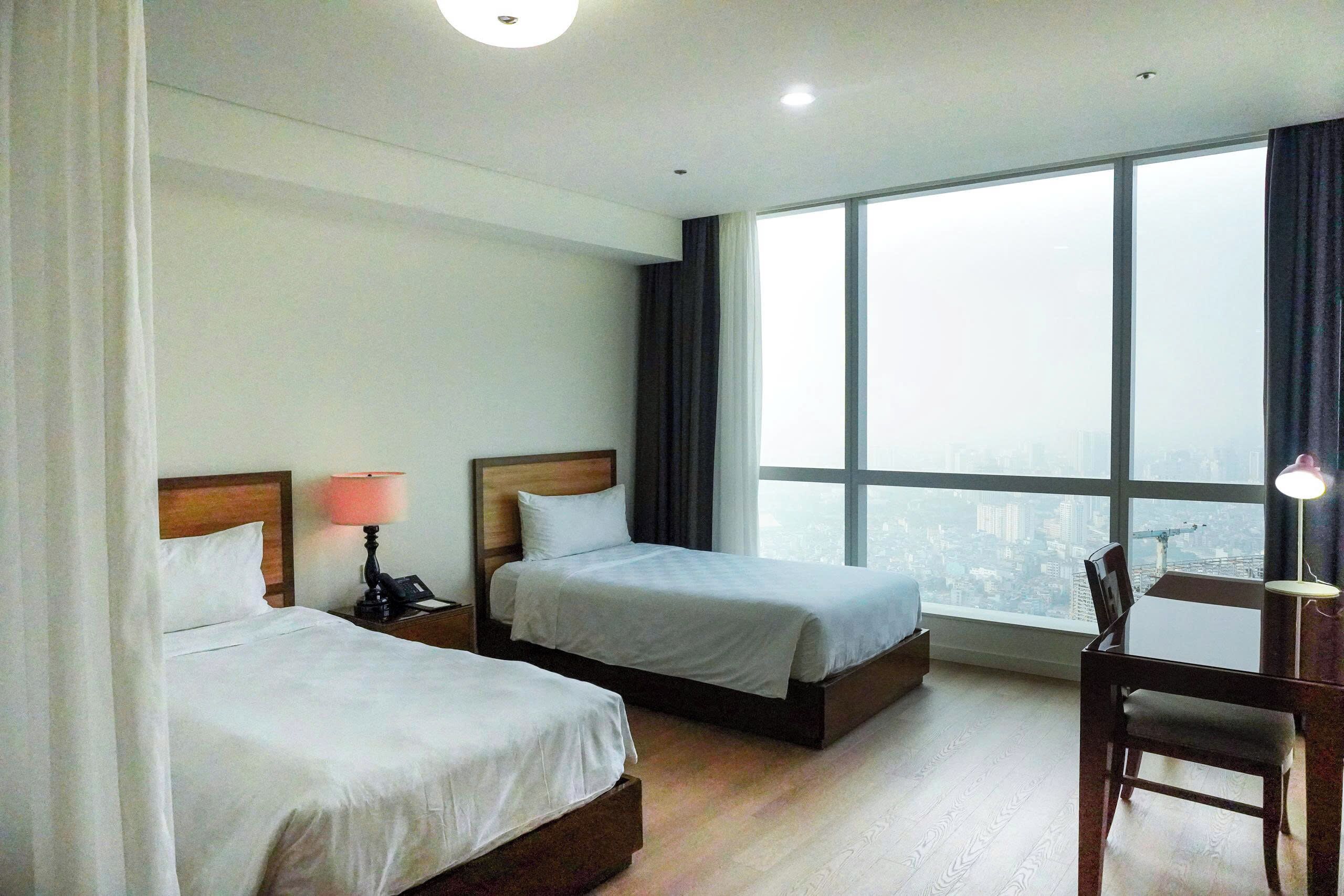 A hotel room with two beds, a desk, and a large window offering a city view.