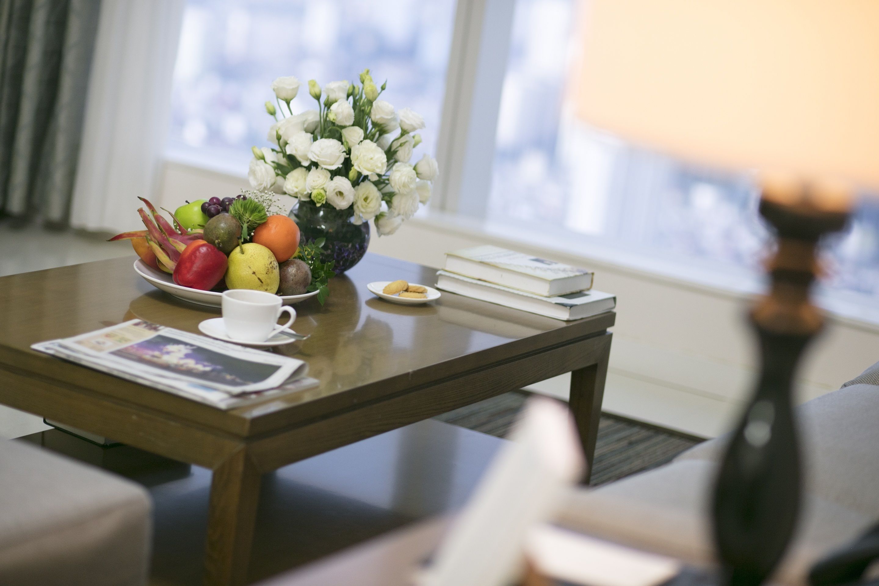 A dining table with a fruit bowl, a vase of flowers, a cup, a plate with cookies, and a book.