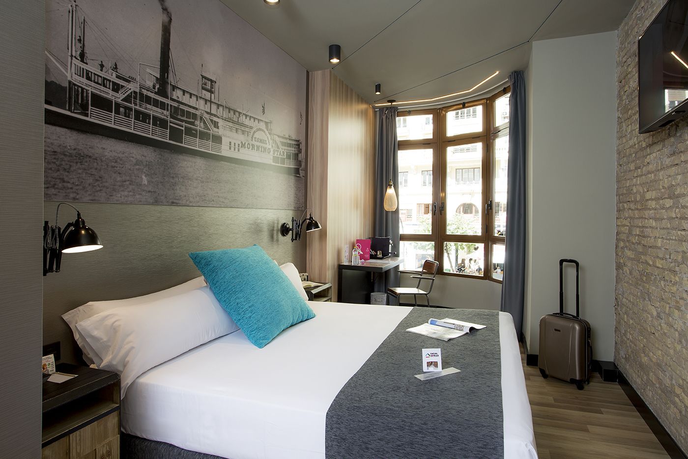 Ein modernes Hotelzimmer mit einem großen Bett, einem Schreibtisch, einem Koffer und einem Wandgemälde.