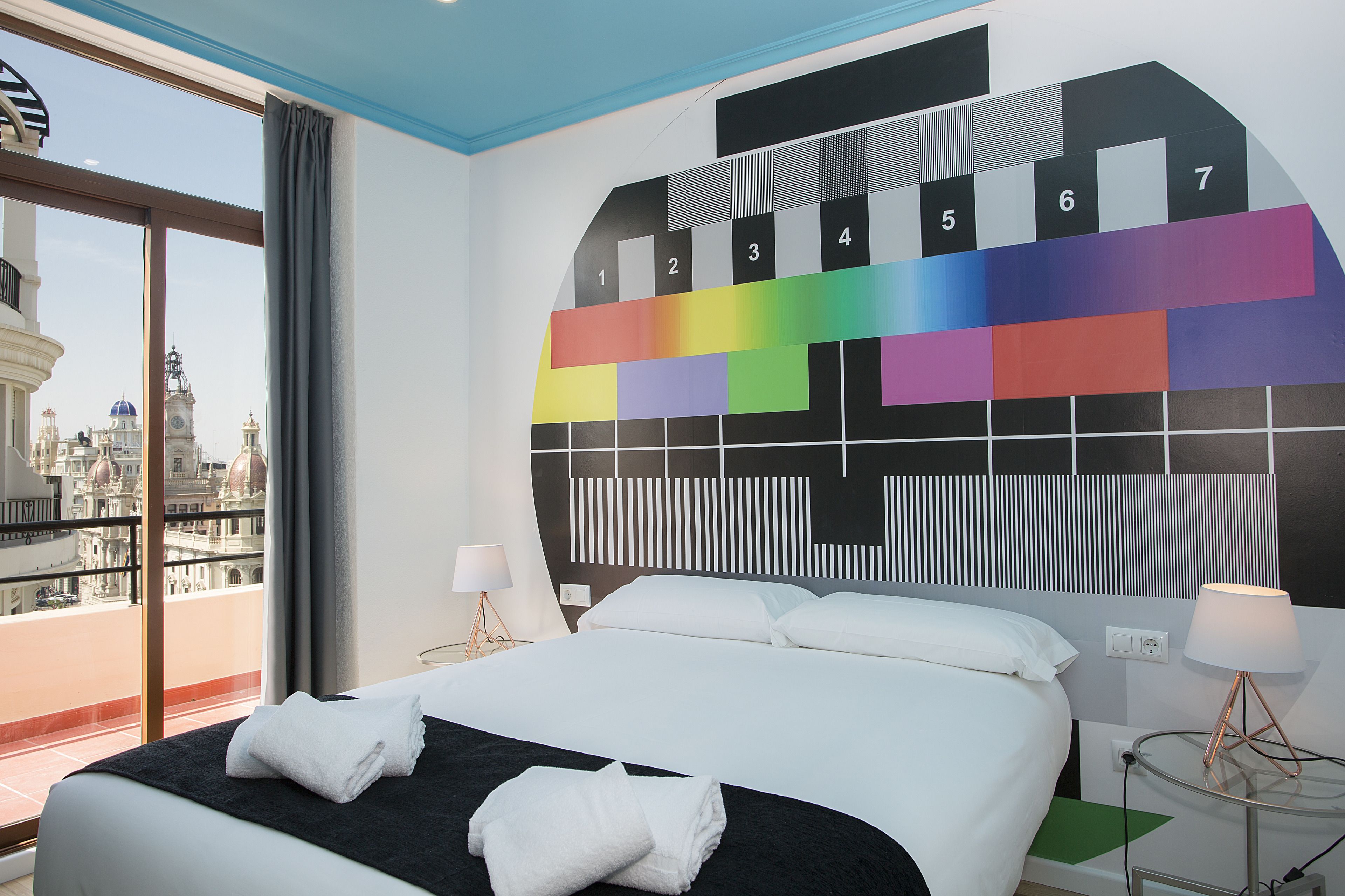 Habitación de hotel con una cama grande, dos mesitas de noche con lámparas, vista a la ciudad desde un balcón y una obra de arte de colores en la pared.