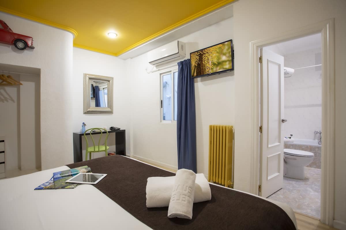 Une chambre d’hôtel avec un lit, un bureau, une chaise, une fenêtre avec des rideaux, un appareil de climatisation, un tableau et une salle de bain visible à travers une porte ouverte.