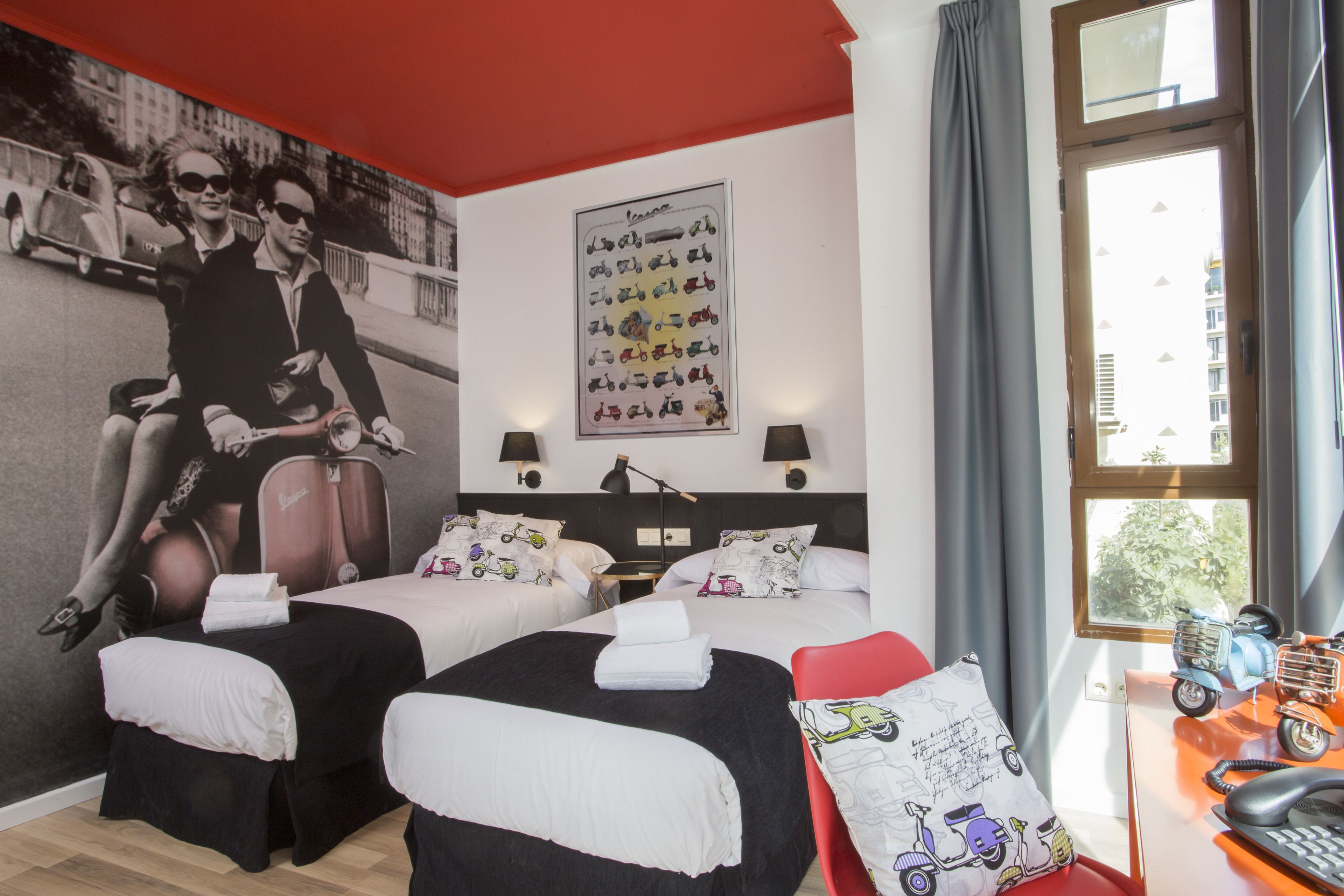 A hotel room with two beds, a red chair, a desk with a phone, and a wall with a vintage car poster.