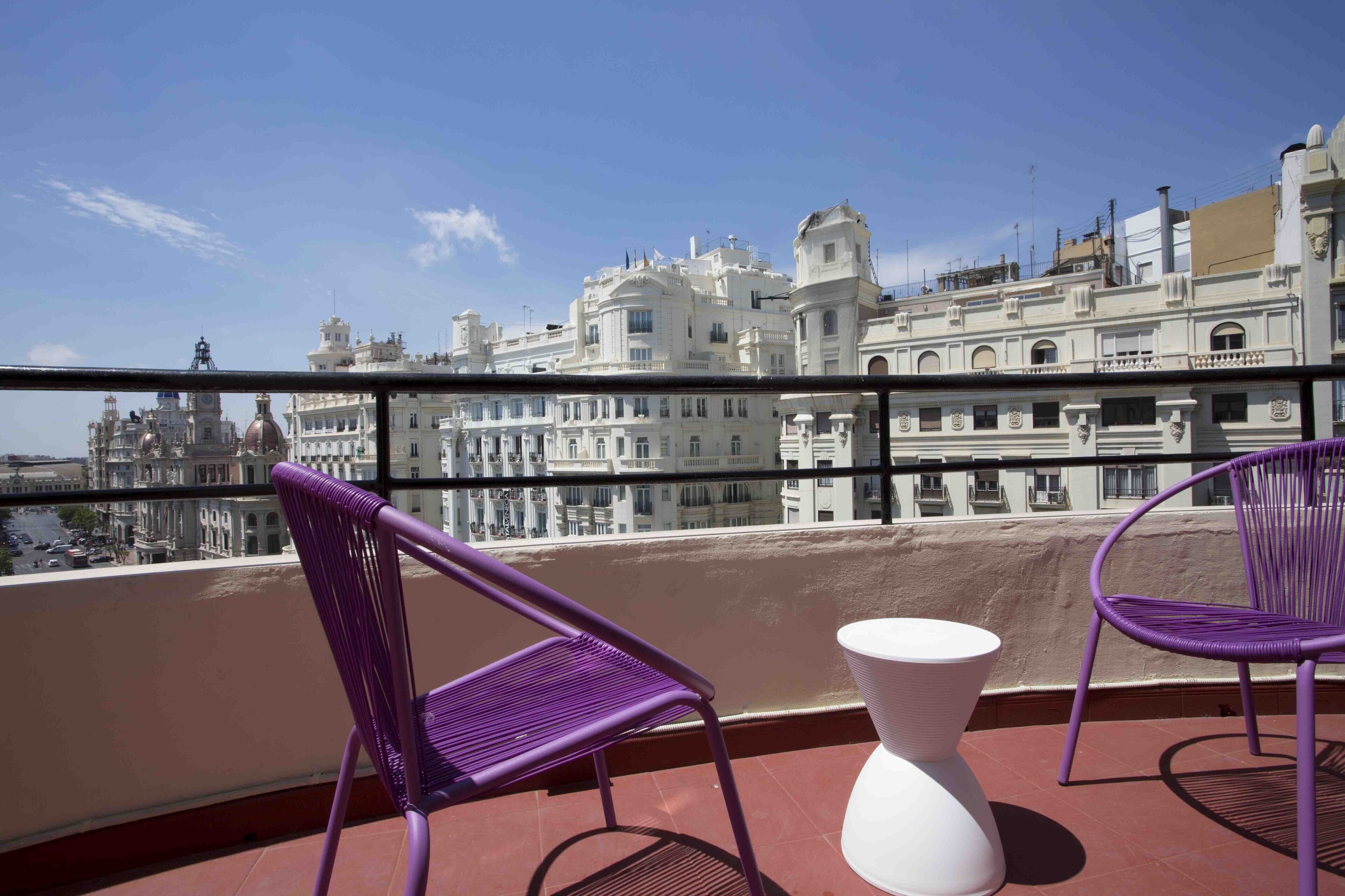 A balcony with two purple chairs and a white table, overlooking a cityscape with historic buildings.