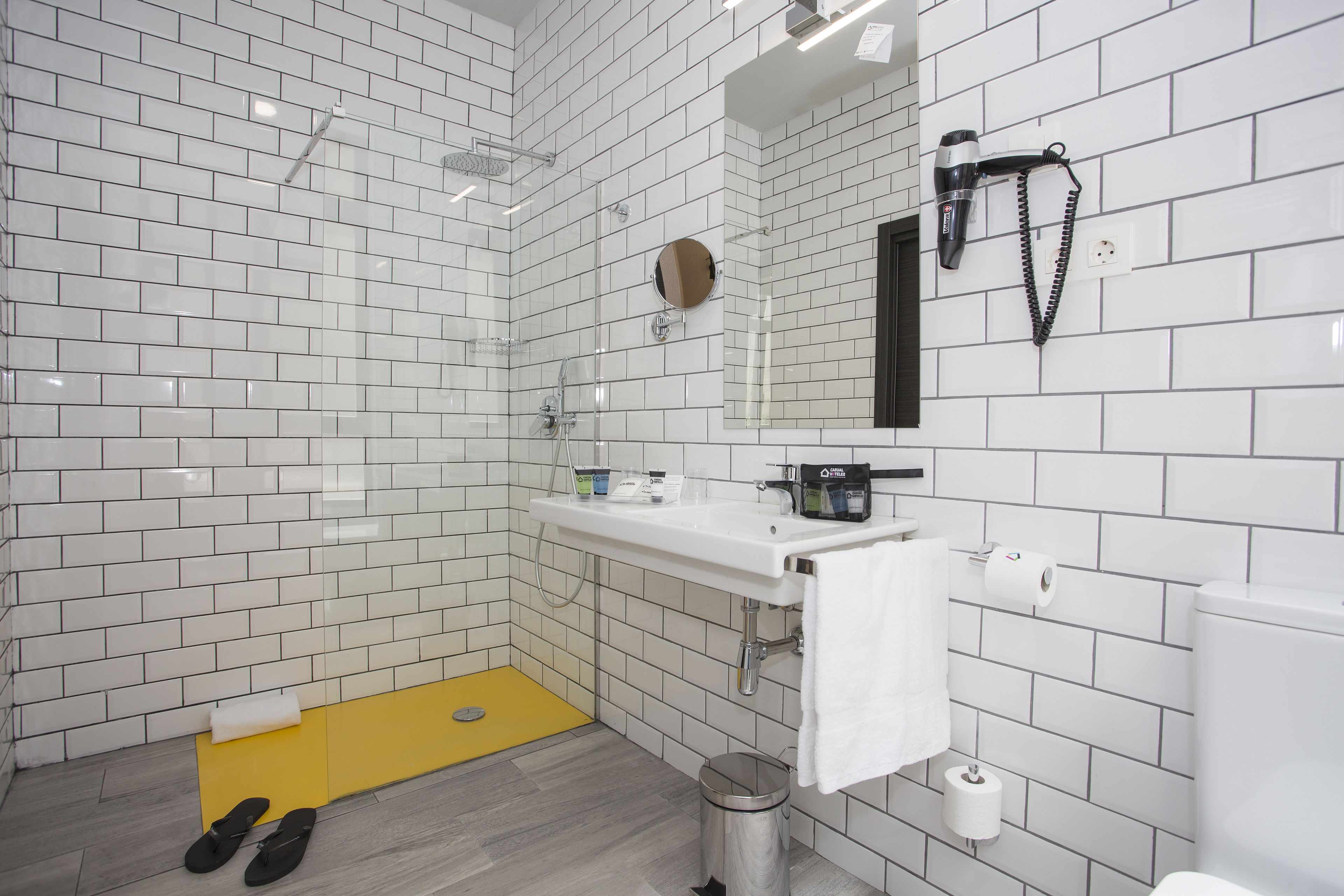 Ein modernes Badezimmer mit weißen Fliesen im U-Bahn-Stil, einer gelben Badmatte, einem Waschbecken mit Spiegel und einem an der Wand montierten Föhn.