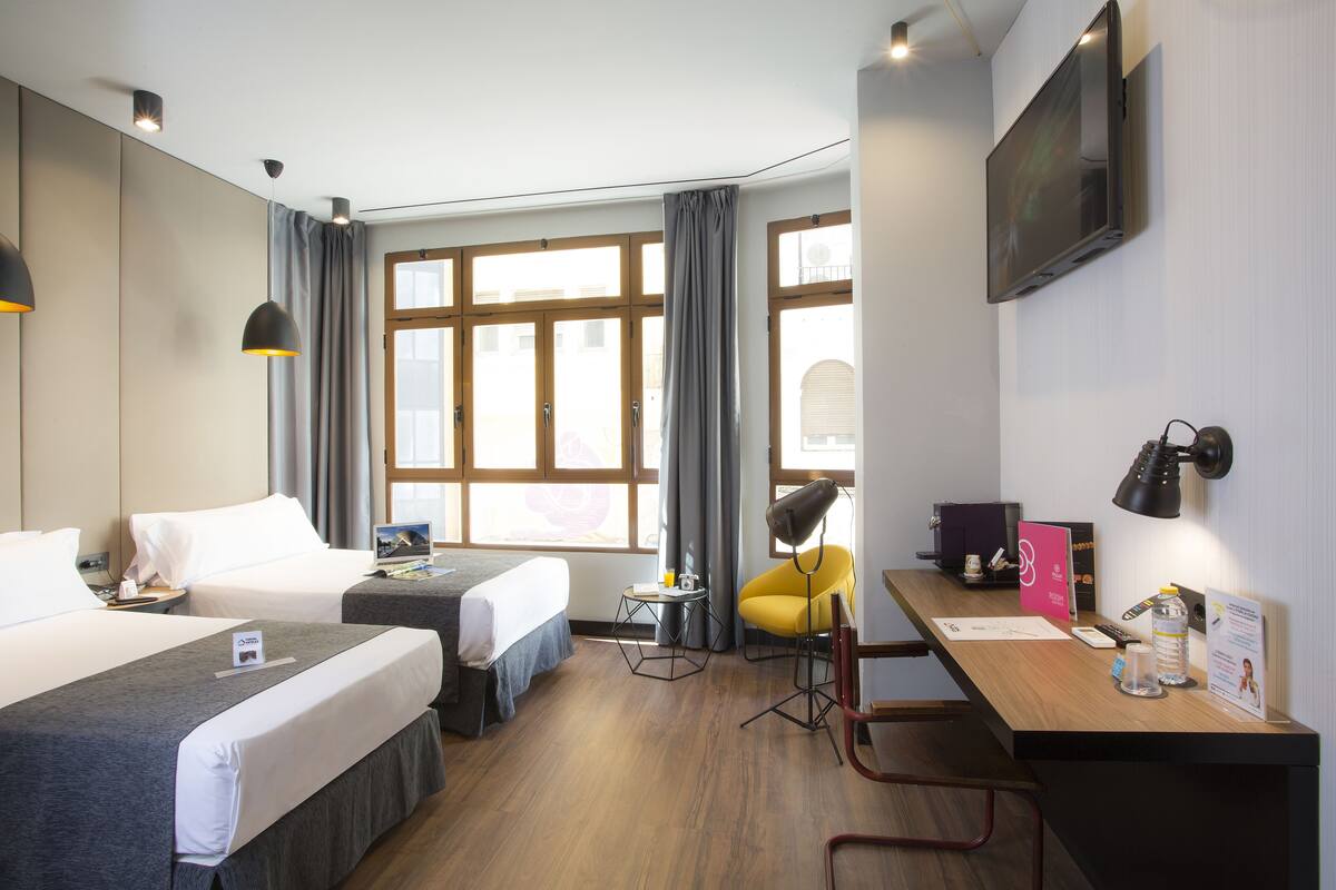 Une chambre d’hôtel avec deux lits, un bureau, une chaise, une télévision et une fenêtre avec des rideaux.