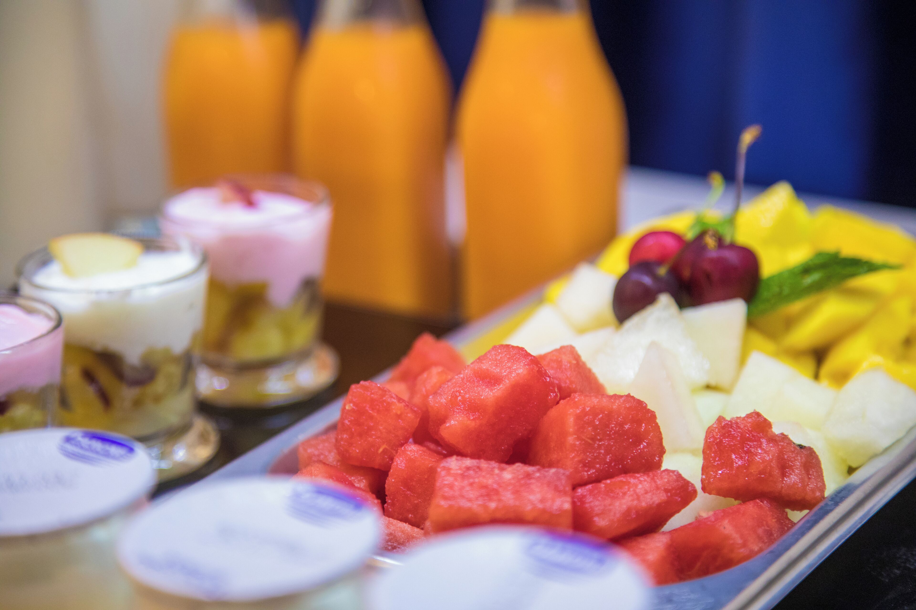 A variety of fruits and beverages on a tray, including watermelon, pineapple, and grapes, with a blurred background of bottles.