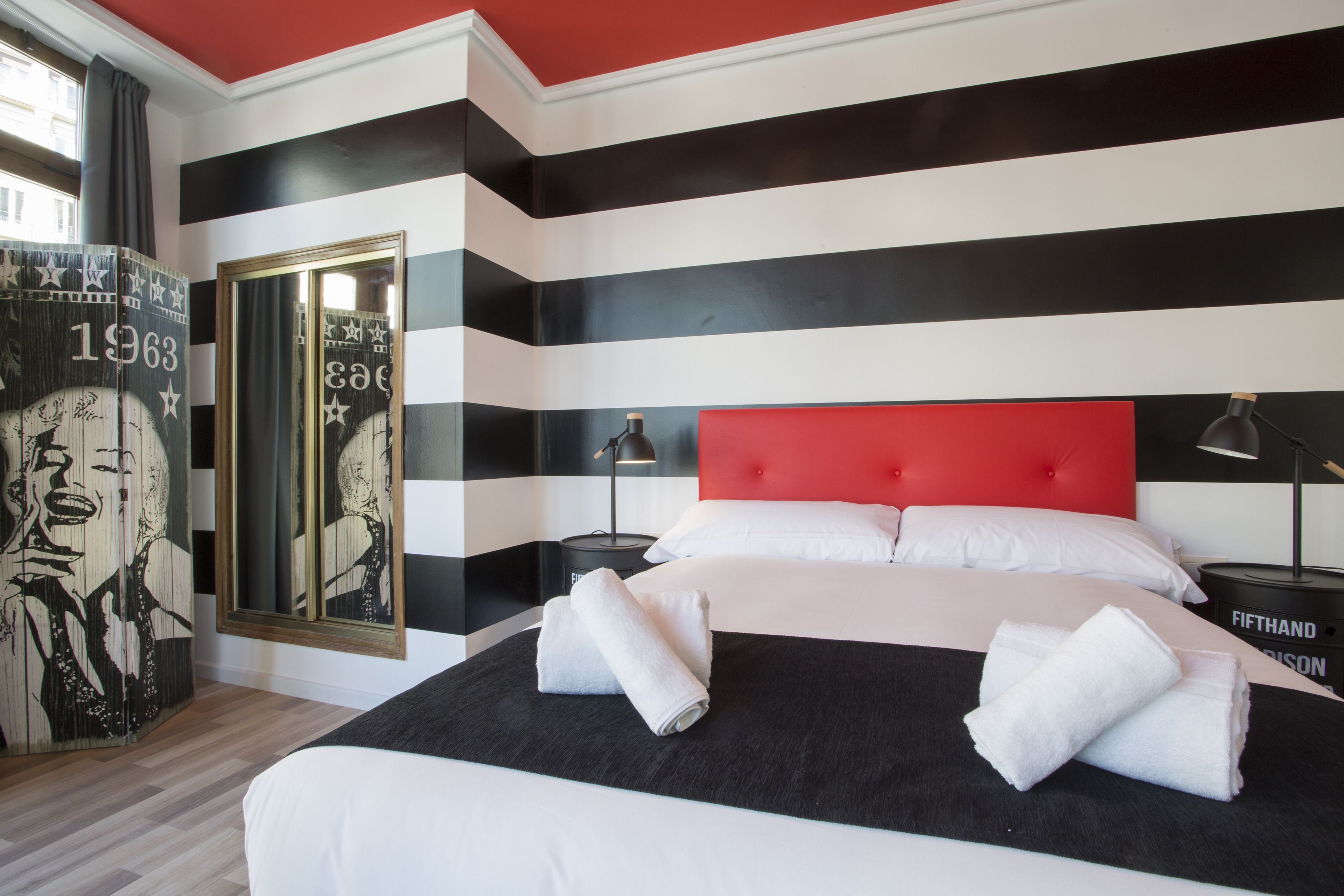 A hotel room with a striped wall design, a red headboard, and a bed with white linens.