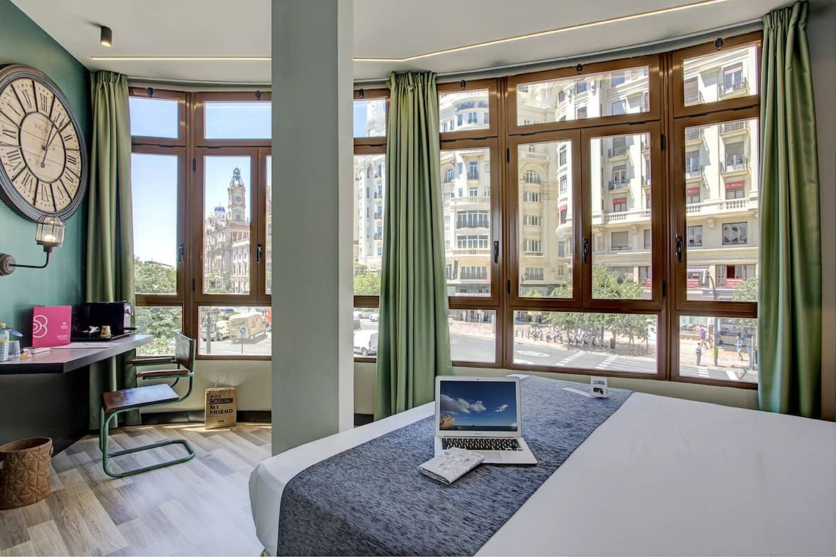 Une chambre d’hôtel moderne dotée de grandes fenêtres, d’un lit, d’un bureau avec un ordinateur portable et offrant une vue sur le paysage urbain.