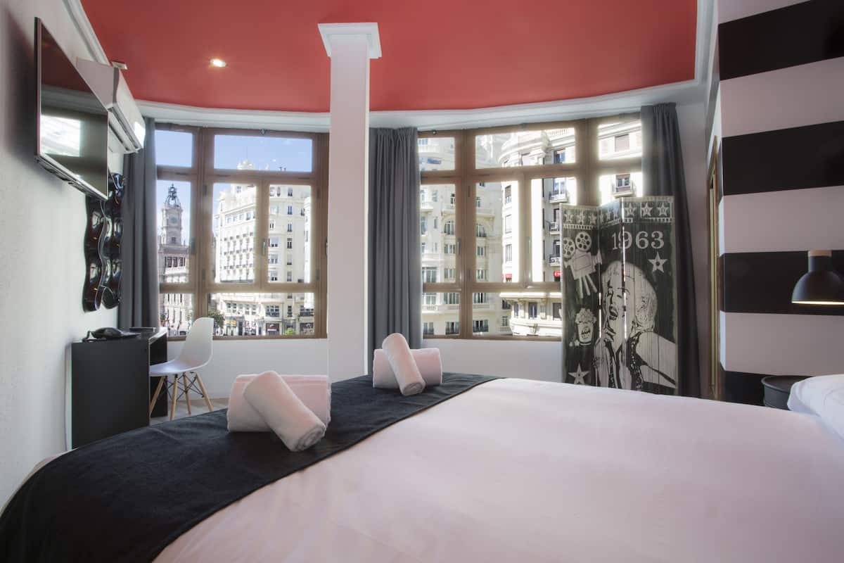 Une chambre d’hôtel avec un grand lit, un bureau, une chaise et une vue sur la ville grâce à de grandes fenêtres.