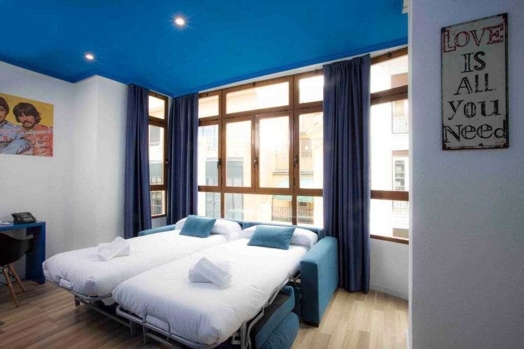 A hotel room with two beds, a desk, and a large window with curtains.