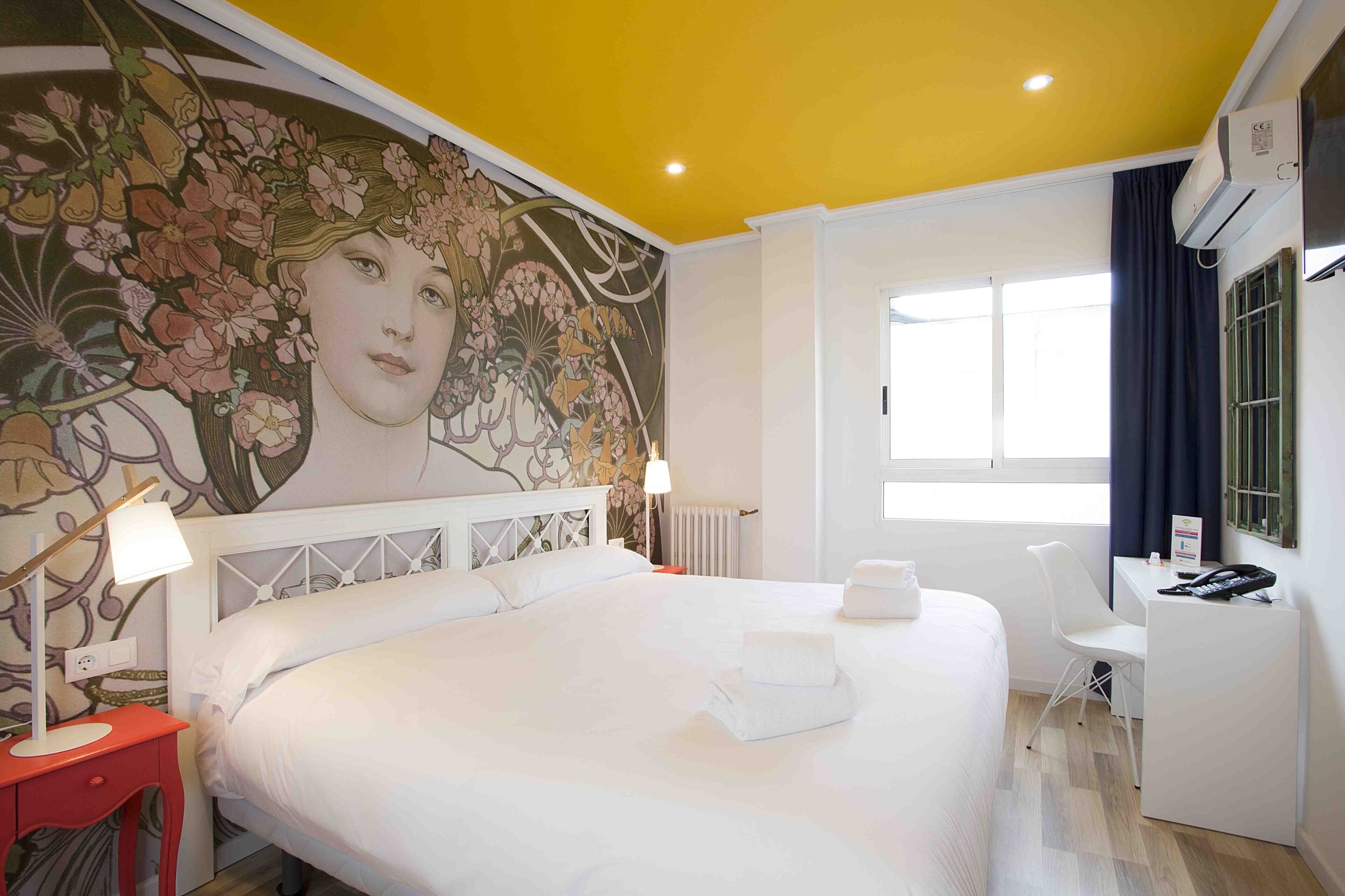 A hotel room with a large bed, a desk, a chair, a radiator, and a window with curtains.