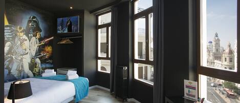 Una habitación de hotel moderna con una cama grande, un escritorio, vistas de la ciudad a través de amplios ventanales y un mural en la pared.