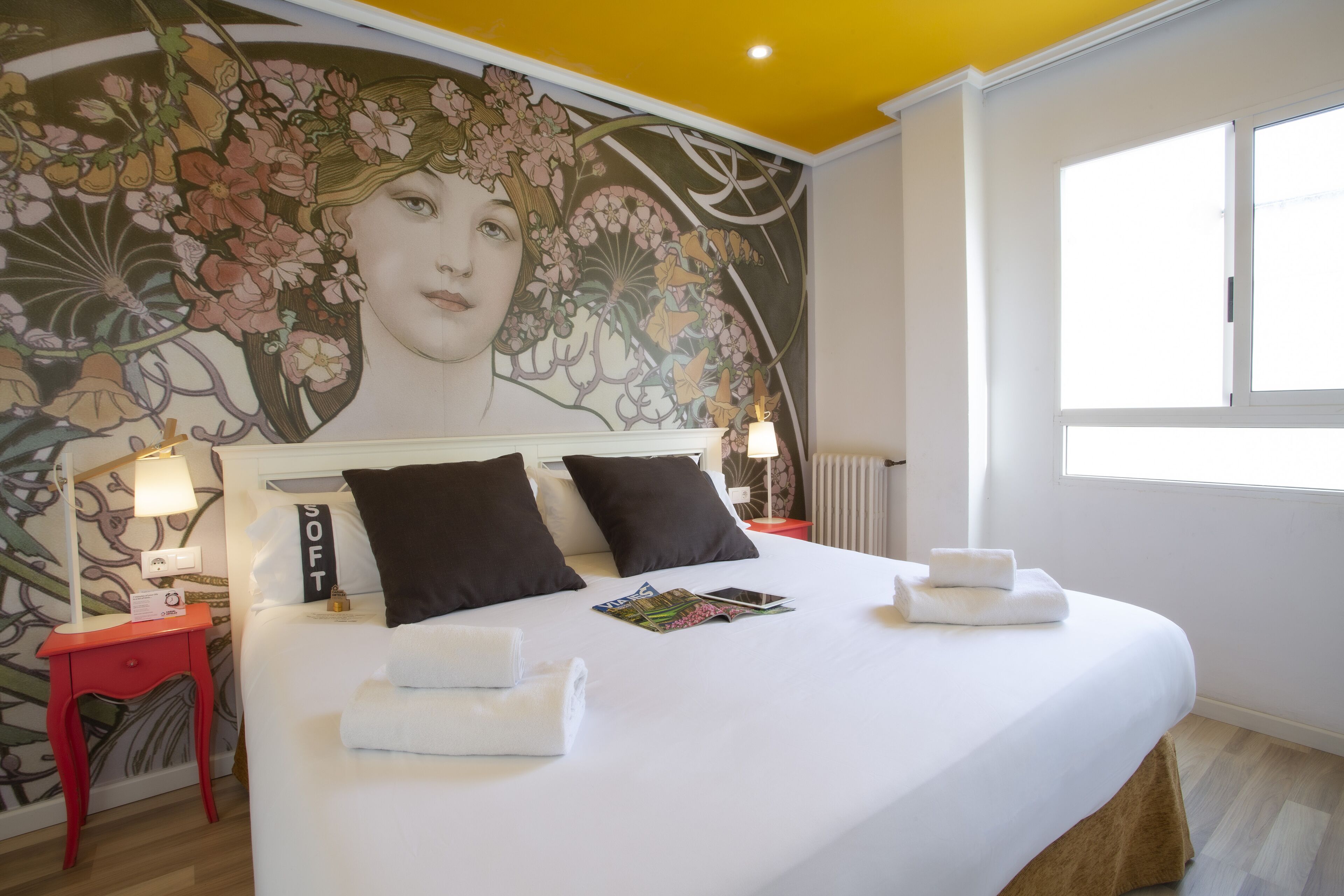 Une chambre d’hôtel avec un grand lit, deux oreillers, une table de chevet avec une lampe, une petite table d’appoint rouge et une fresque florale sur le mur.