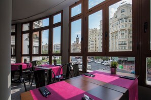 En restaurang med stora fönster, bord med rosa dukar och utsikt över stadens byggnader.
