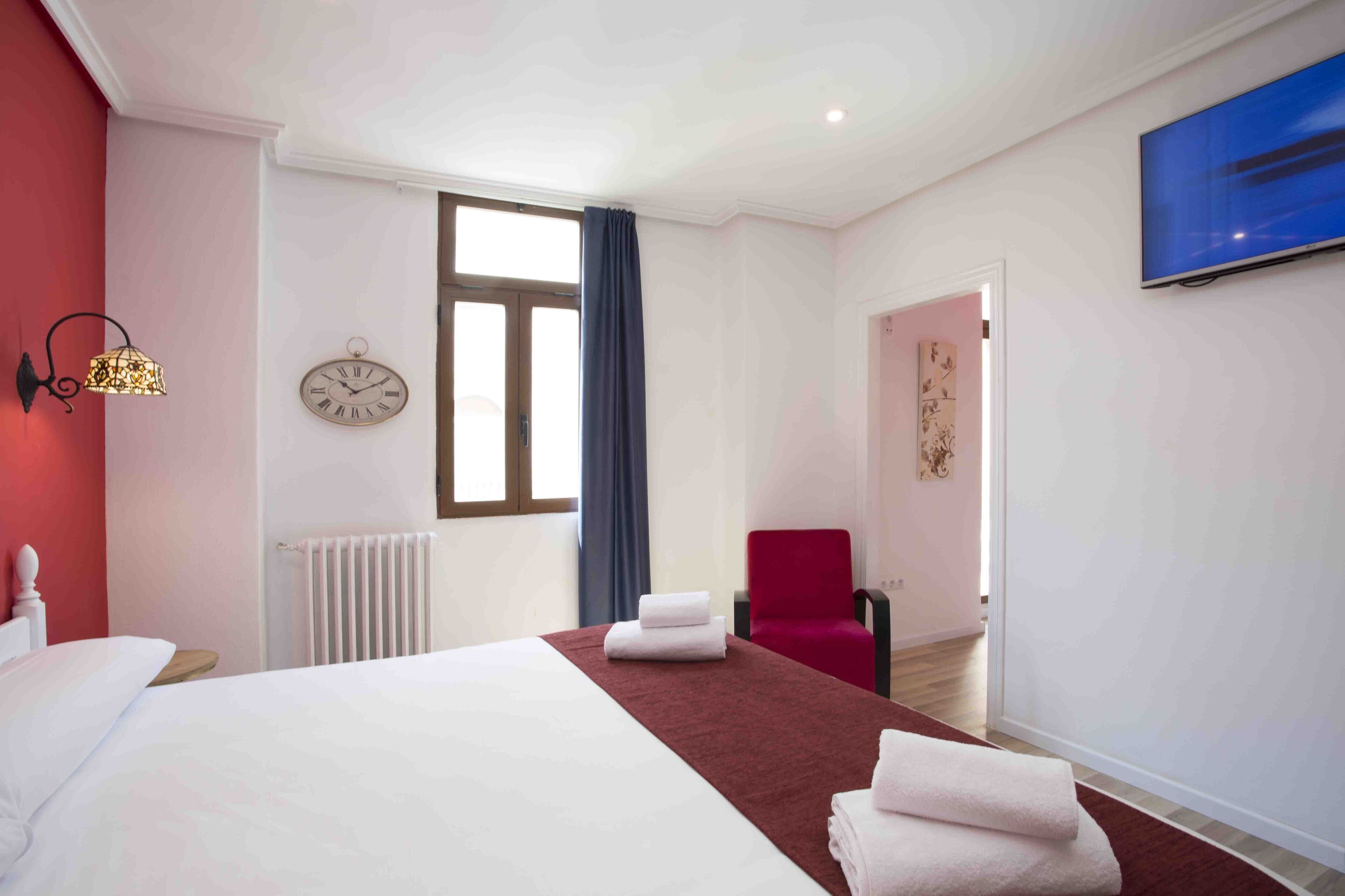 A hotel room with a large bed, a red chair, a clock, and a TV on the wall.