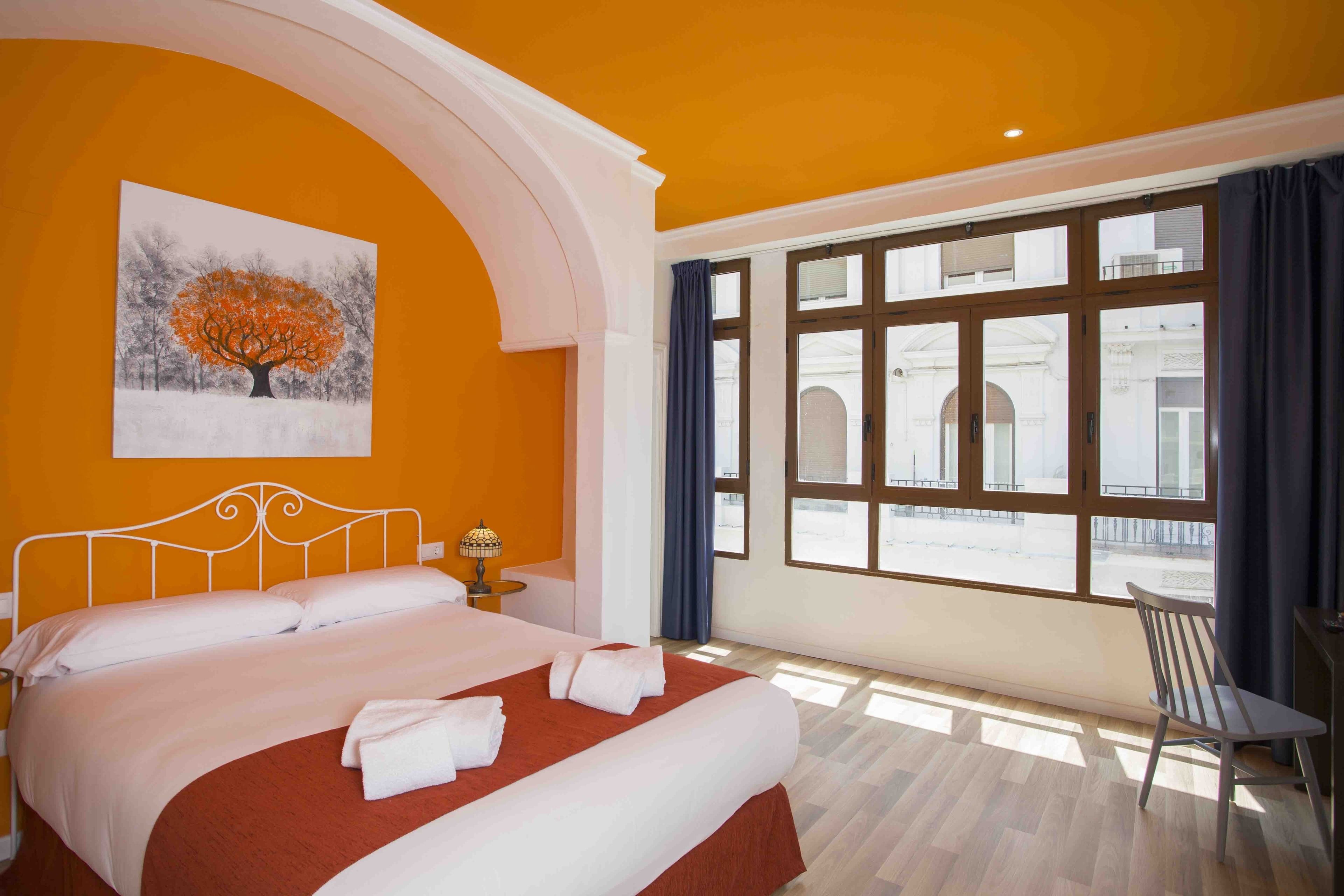 Une chambre d’hôtel avec un grand lit, un tableau représentant un arbre, une fenêtre avec des rideaux et une vue sur un bâtiment à l’extérieur.
