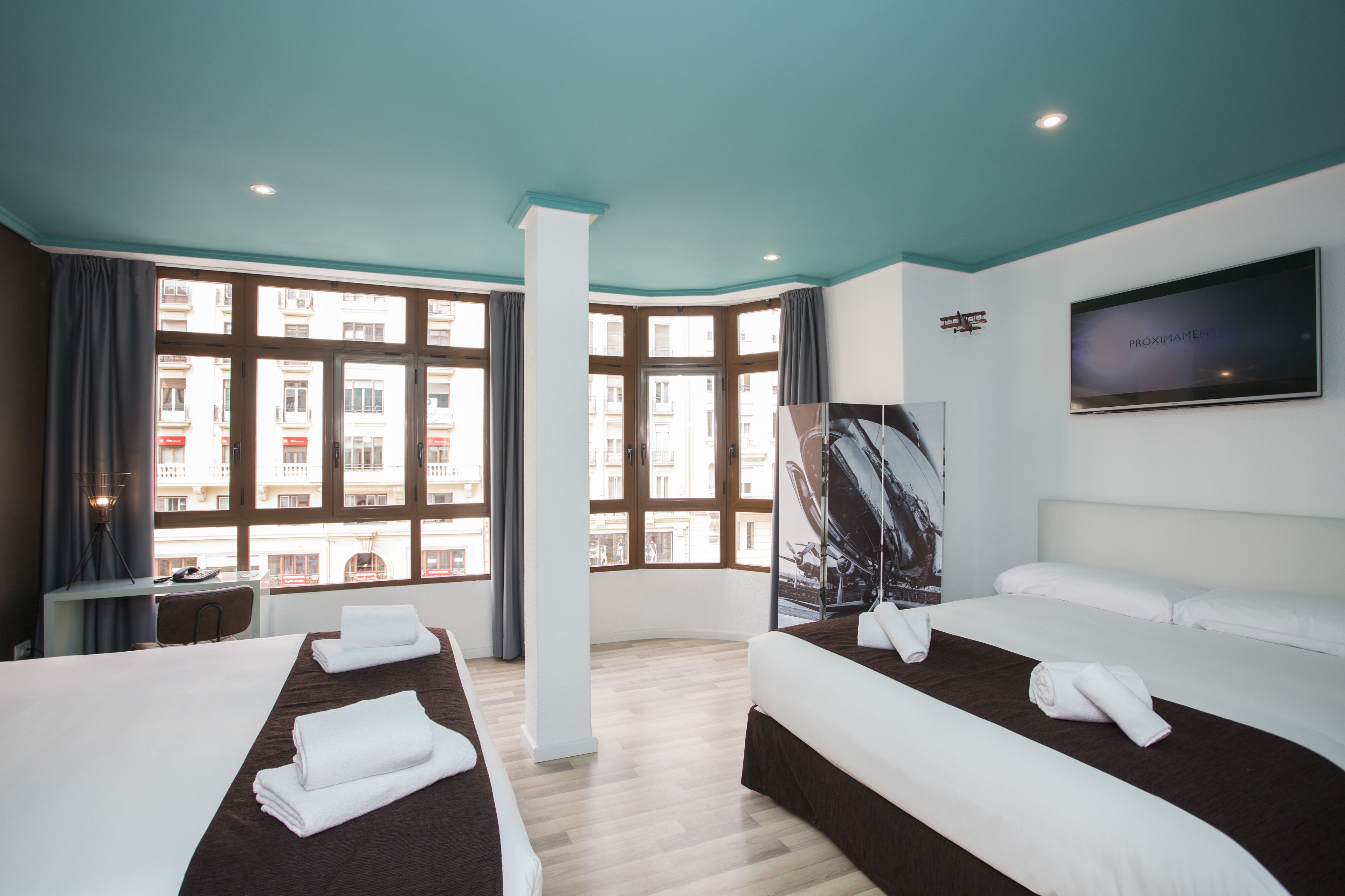 A hotel room with two beds, a desk, and a large window with a view of buildings.