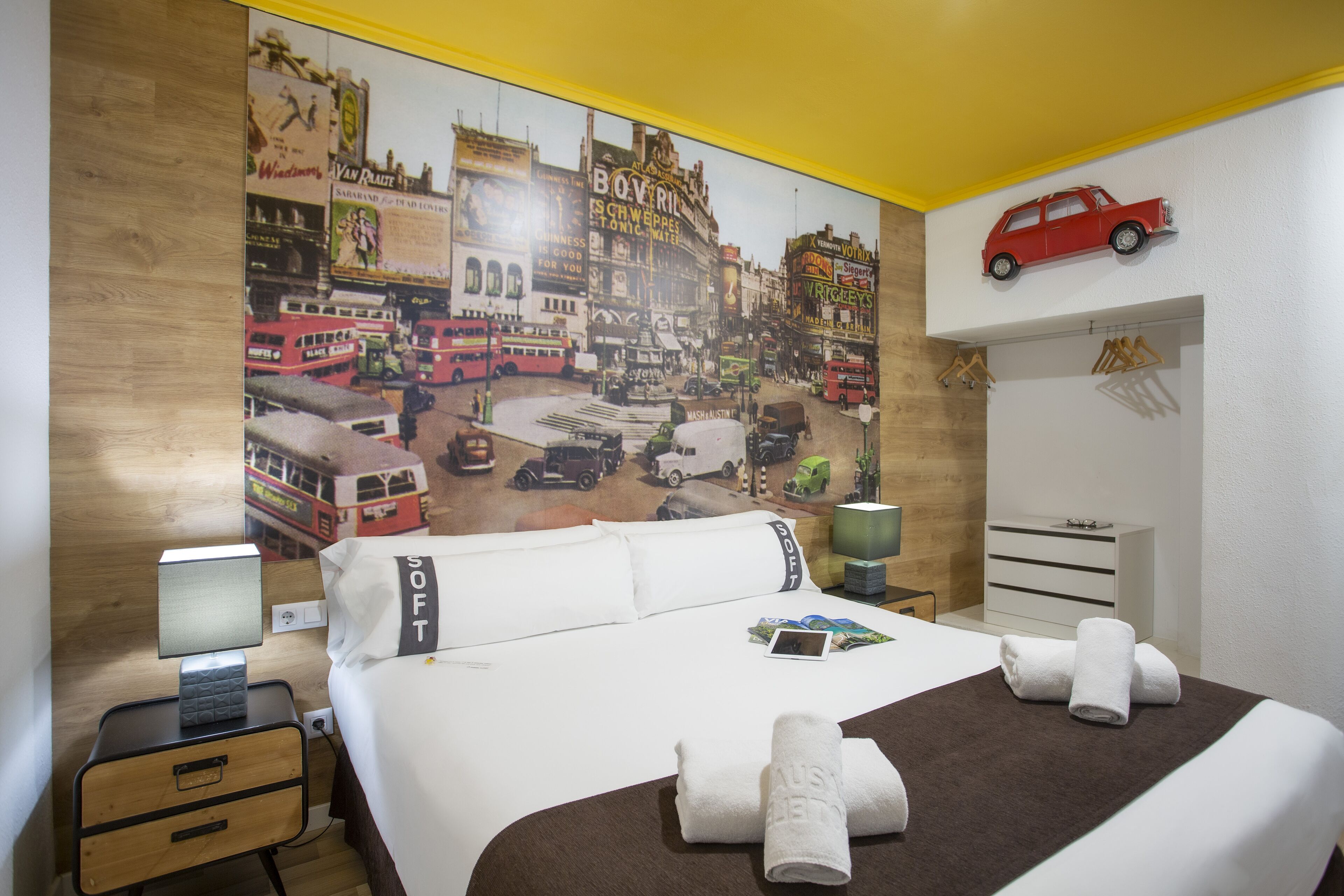 A hotel room with a large cityscape mural, a bed with white linens, a nightstand with a lamp, a dresser, and a red toy car on the wall.