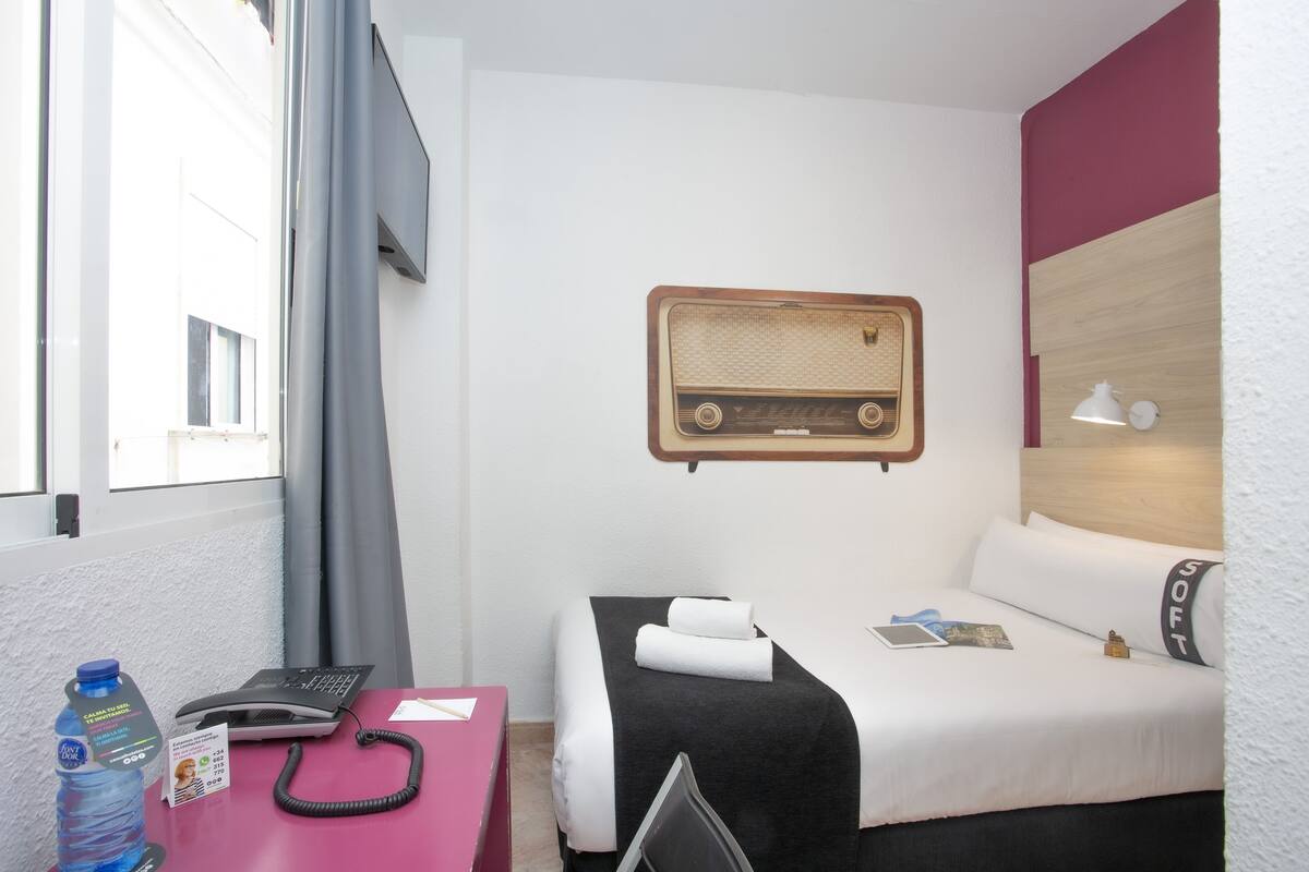 Une chambre d’hôtel avec un lit, un bureau avec un ordinateur portable, un téléphone, une bouteille d’eau et une radio fixée au mur.