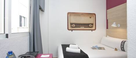 A hotel room with a bed, a desk with a laptop, a telephone, a water bottle, and a radio on the wall.