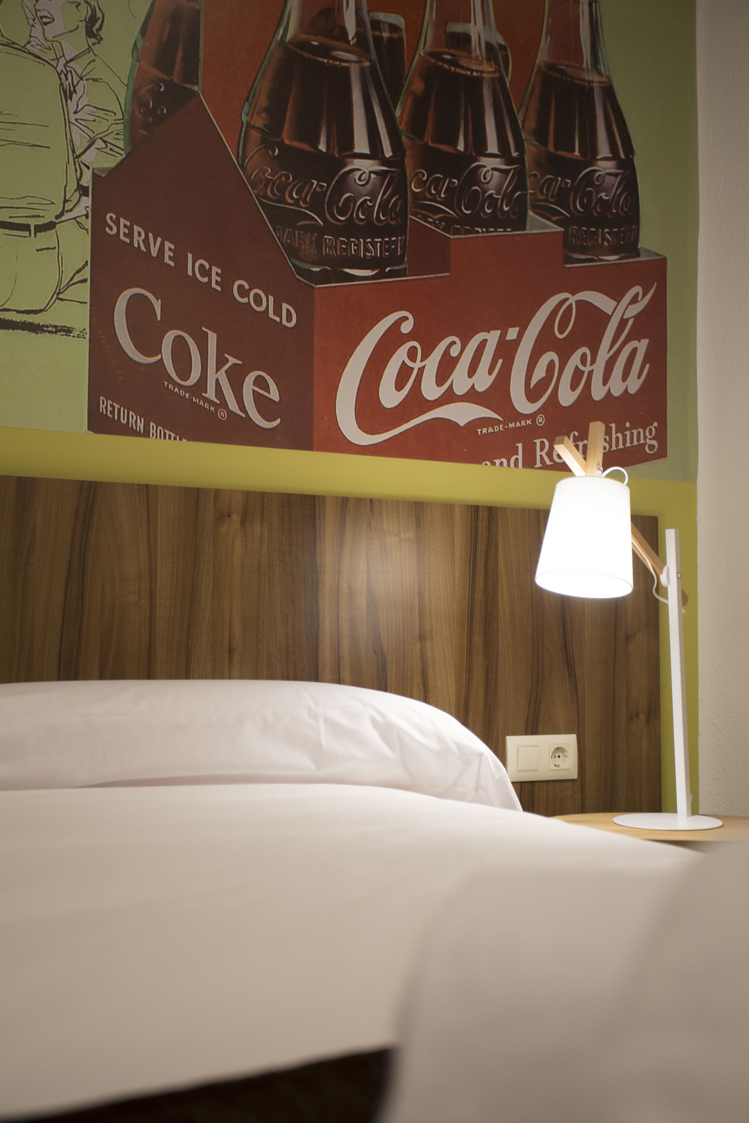 A hotel room with a bed, a nightstand, a lamp, and a wall with a Coca-Cola advertisement.