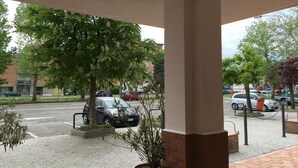 A covered outdoor area with a brick column, potted plants, and a view of parked cars and buildings in the background. - Hotel Commercio (Campogalliano)