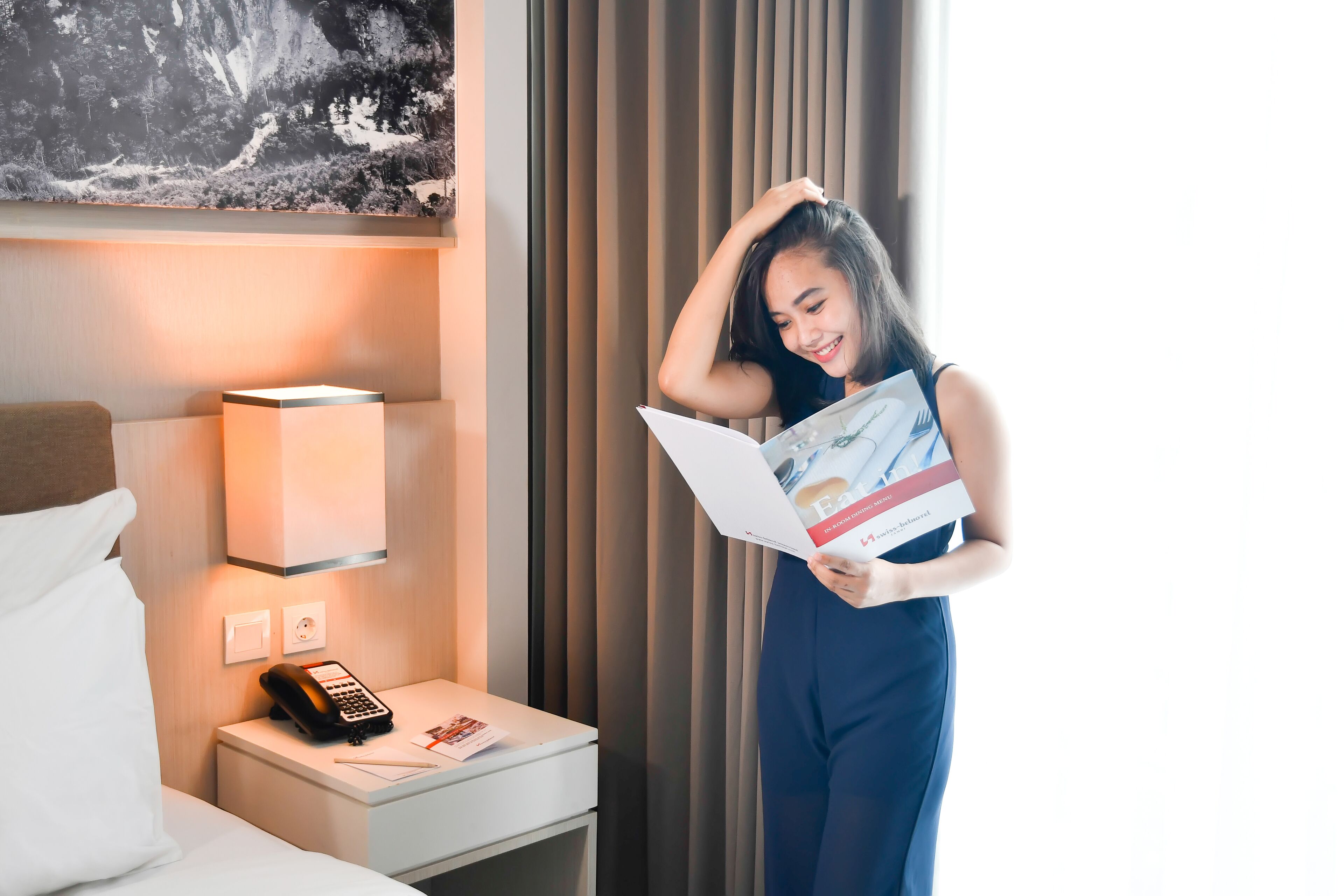 Et hotelværelse med en seng, et natbord med en telefon, en lampe og et billede på væggen.