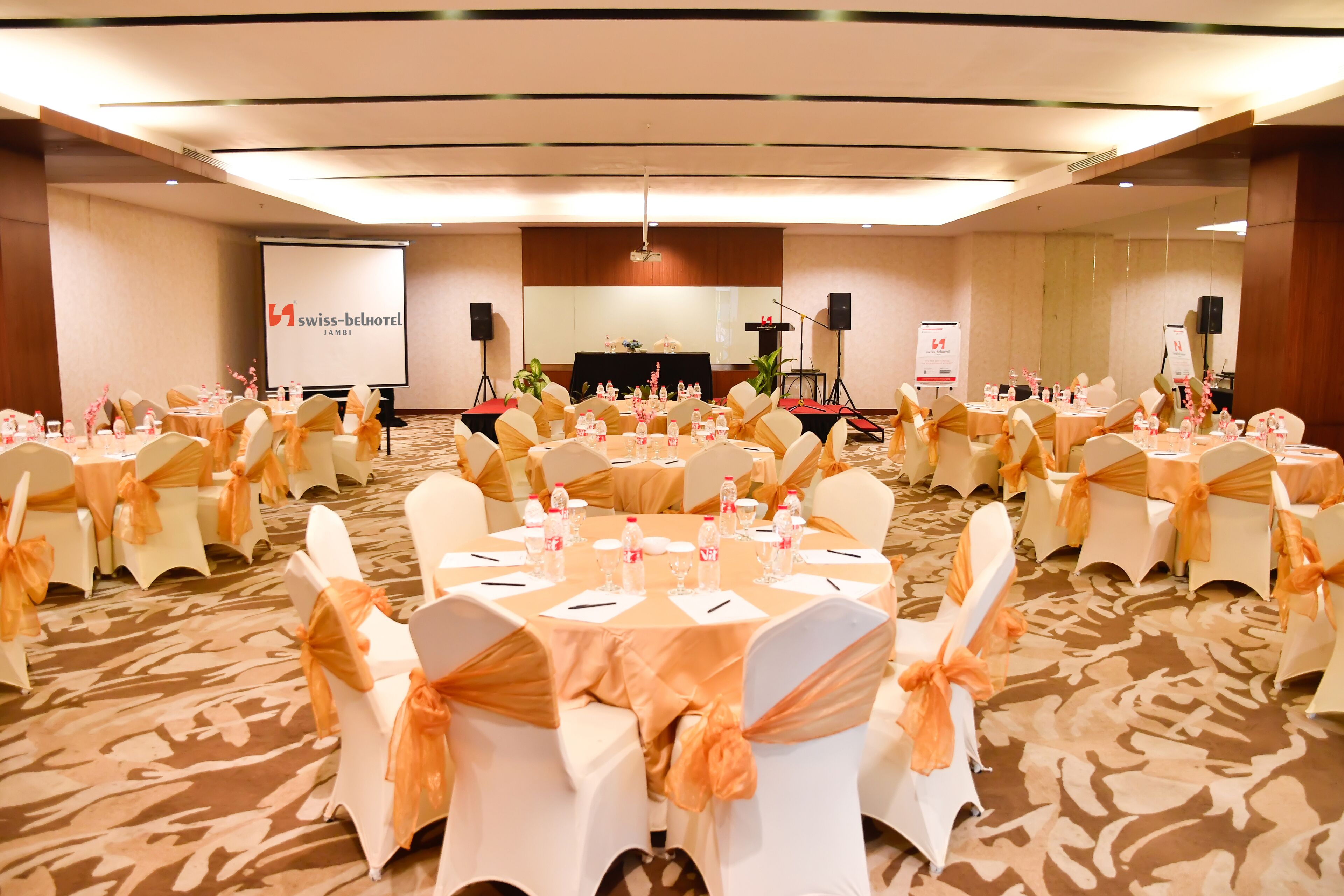 Ein Konferenzraum mit runden Tischen, weißen Stühlen mit orangefarbenen Bändern und einer Projektionsleinwand, auf der "swiss-belhotel" zu sehen ist.