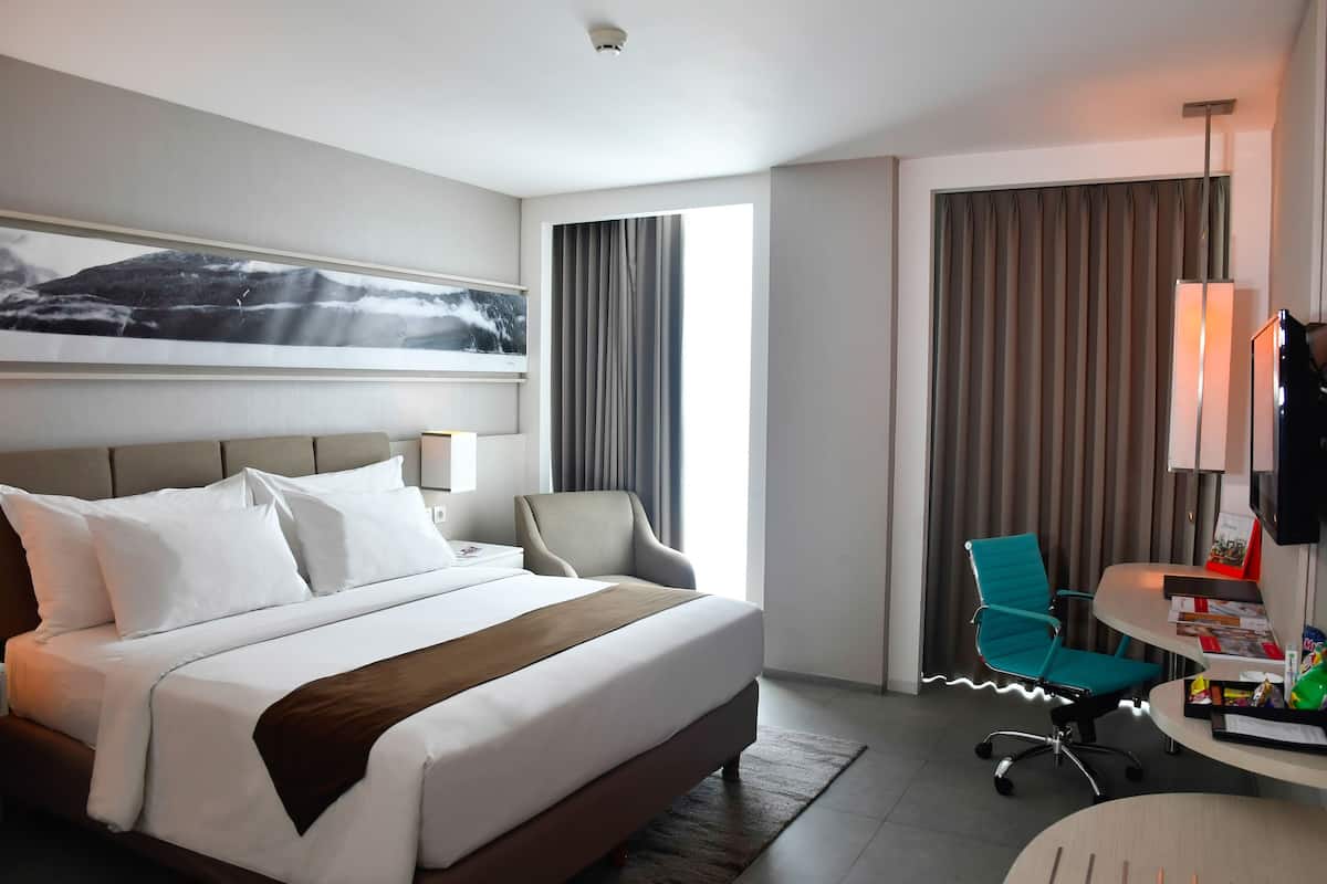 Une chambre d’hôtel avec un lit, un bureau, une chaise, une lampe et une fenêtre avec des rideaux.
