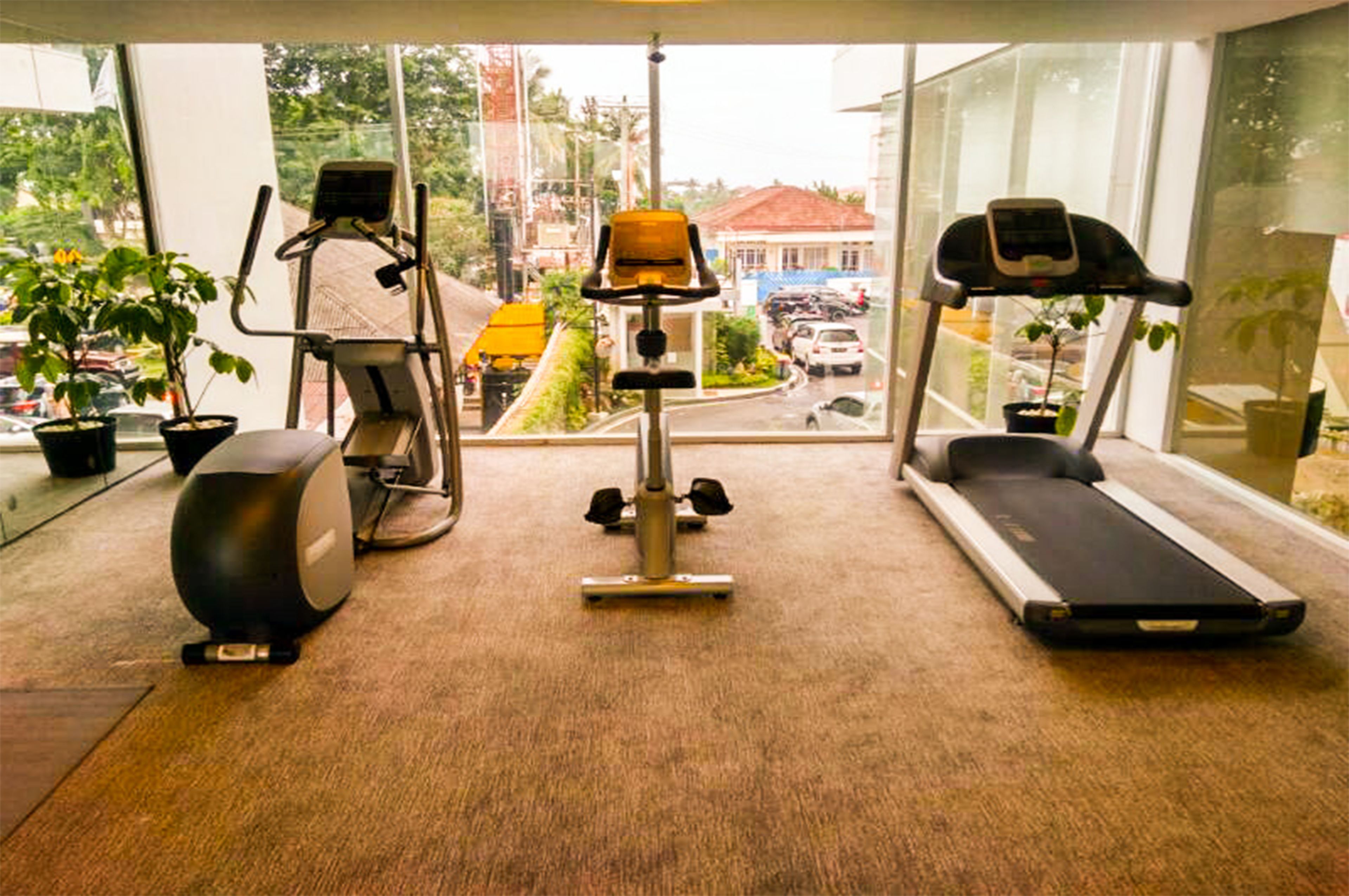 Un gimnasio con equipo de ejercicio, incluyendo cinta de correr, elíptica y bicicleta estática, con ventanas amplias que ofrecen vista a la calle.