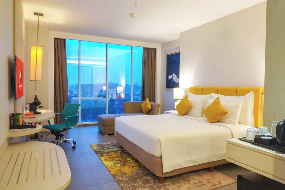 Une chambre d’hôtel avec un grand lit, un bureau, une chaise, une télévision et une vue sur la ville grâce à de grandes fenêtres.