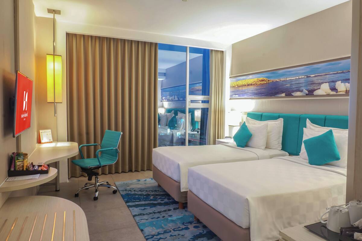 Une chambre d’hôtel avec deux lits, un bureau, une chaise, une télévision et une grande fenêtre avec des rideaux.
