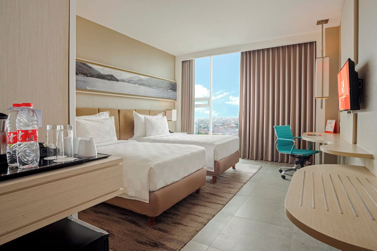 Une chambre d’hôtel avec deux lits, un bureau, une chaise, une télévision et une grande fenêtre donnant sur la ville.