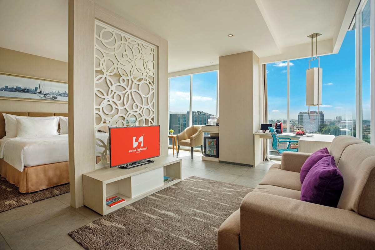 Une chambre d’hôtel moderne équipée d’un lit, d’une télévision, d’un bureau et d’une grande fenêtre donnant sur la ville.