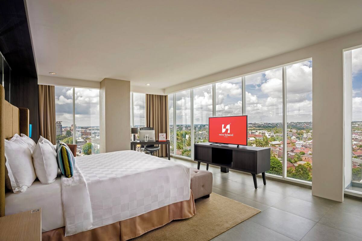 Une chambre d’hôtel moderne dotée d’un grand lit, d’un bureau avec un ordinateur, d’une télévision et offrant une vue sur la ville grâce à de grandes fenêtres.