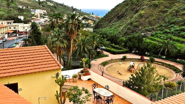 A residential area with a round playground, a yellow building with a tiled roof, and a view of a hillside with vegetation and a road. - Hotel Rural Villa Hermigua (Hermigua)