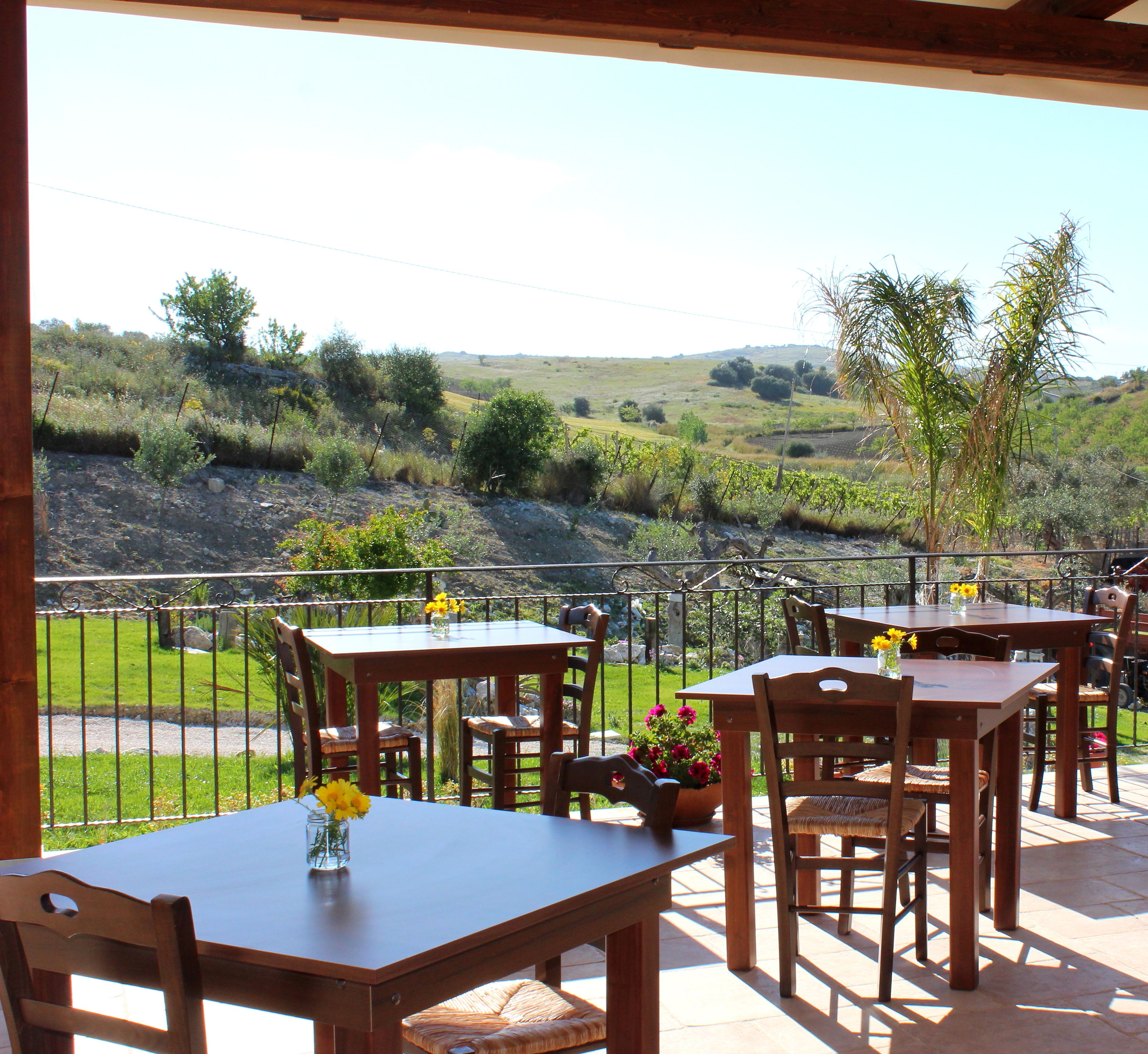 An outdoor patio with wooden tables and chairs, a view of a hilly landscape, and a metal fence.
