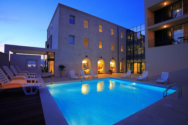 A swimming pool with lounge chairs, a building with multiple windows and balconies, and a clear sky. - Hotel Lipa (Postira)