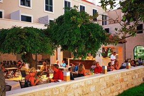 An outdoor dining area with stone walls and tables, surrounded by greenery and a building in the background. - Hotel Lipa (Postira)