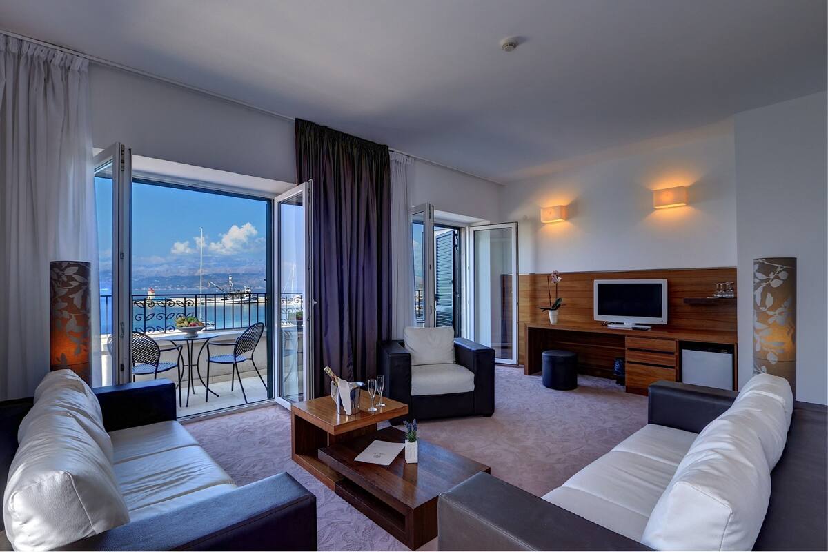 A modern hotel room with a balcony, a sofa, a TV, and a dining area with a table and chairs.