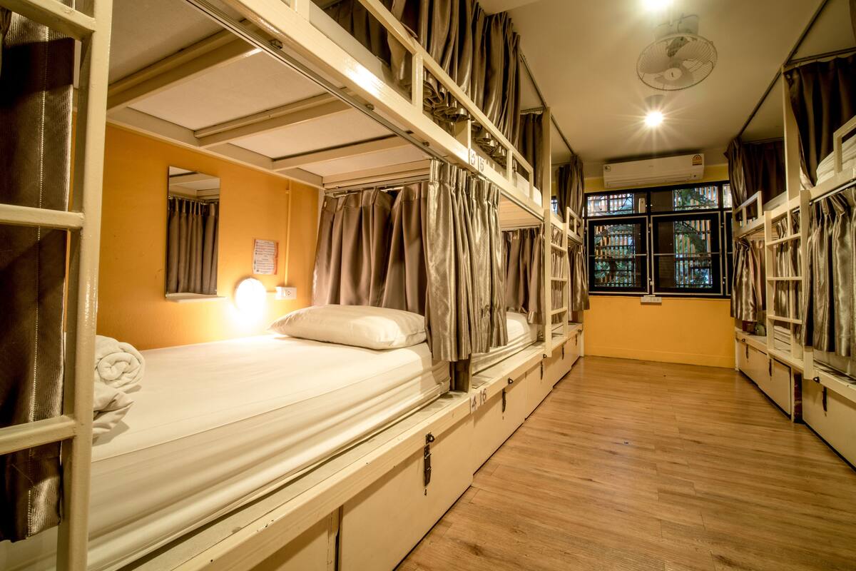 A dormitory room with bunk beds, a ceiling fan, a window with curtains, and a bedside lamp.