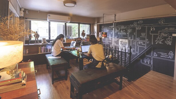 A cozy workspace with two individuals at desks, a large window, and a wall with a chalkboard design. - Bharani Hostel (Bangkok)