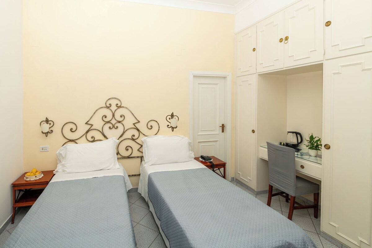 A room with two beds, a desk, a chair, a wardrobe, and a small table with a bowl of fruit.
