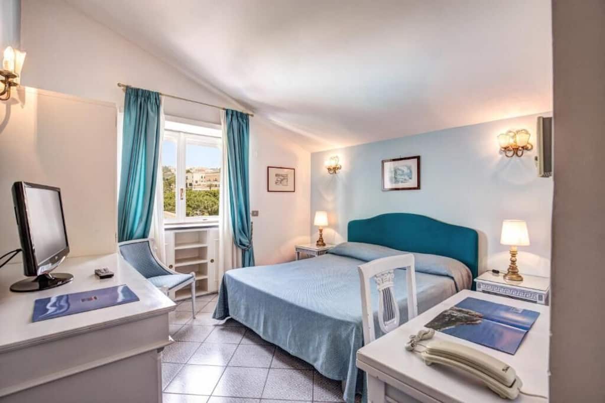 Habitación de hotel con cama, mesitas de noche, escritorio con computadora, silla y ventana con cortinas.