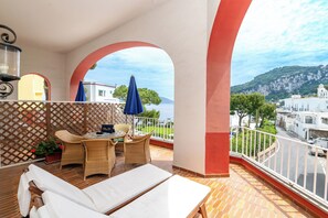 Un balcón con una silla lounge blanca, mobiliario de mimbre y vistas al mar y edificios.