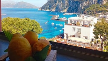 Une vue sur la côte, avec une montagne en arrière-plan, un bâtiment blanc doté de balcons, et un panier de fruits posé sur un rebord de fenêtre.