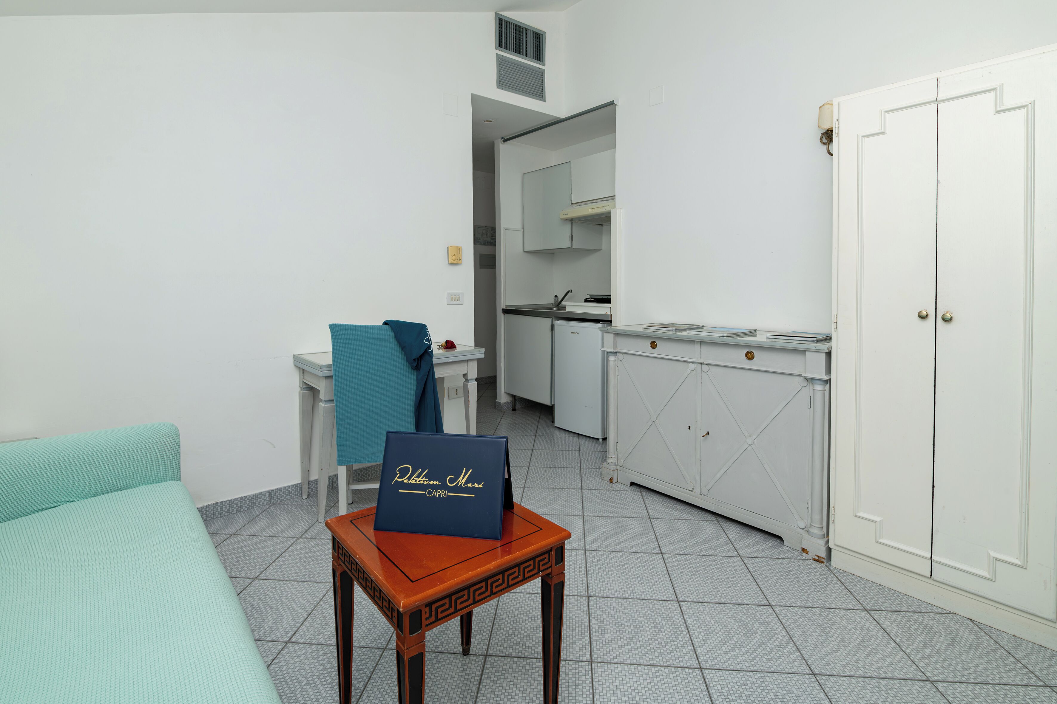 A compact room with a bed, a small table with a sign, a kitchenette, and a wardrobe.