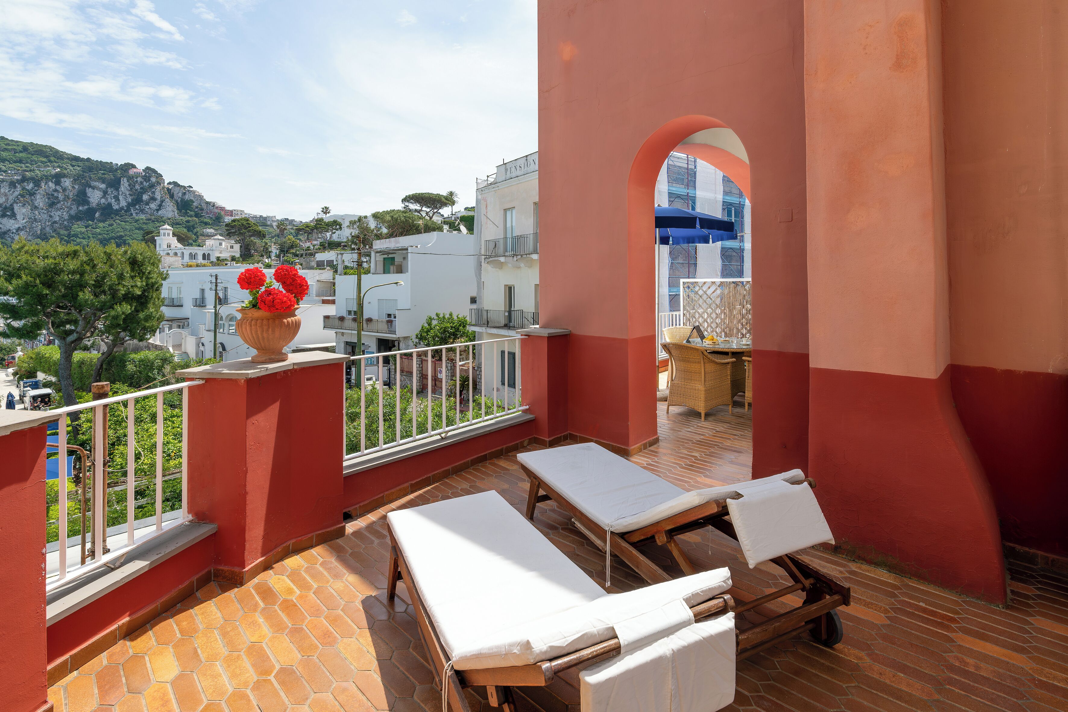 Un balcon avec deux chaises longues, un pot de fleurs orné de fleurs rouges et une vue sur des immeubles et une montagne.